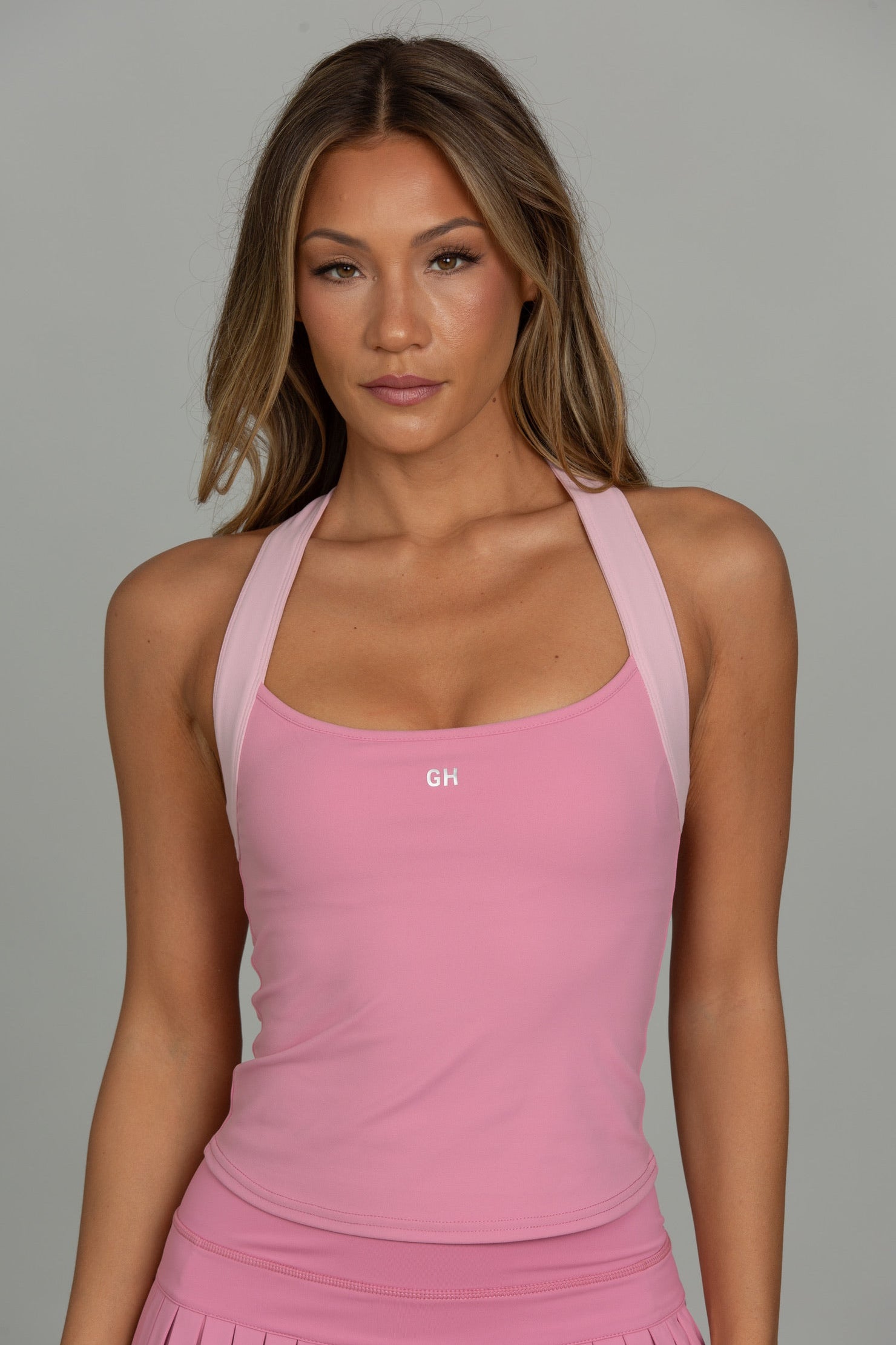 A woman modeling a pink athletic halter tank top with a small white 'GH' logo on the chest, paired with a matching pink pleated skirt, against a solid gray studio background.