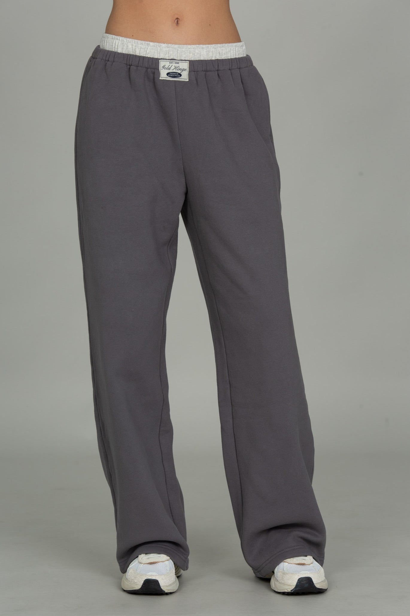 A person shown from the waist down wearing loose gray lounge pants with a white elastic waistband and white sneakers, photographed against a plain light gray studio background.