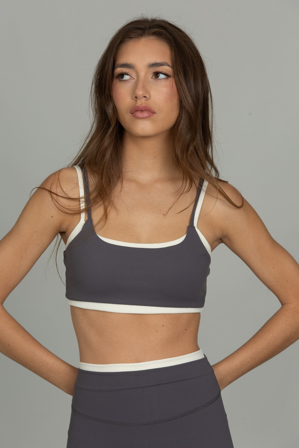 A woman modeling a steel gray double-layer sports bra with contrasting ivory trim, paired with matching gray leggings, against a plain gray studio background.