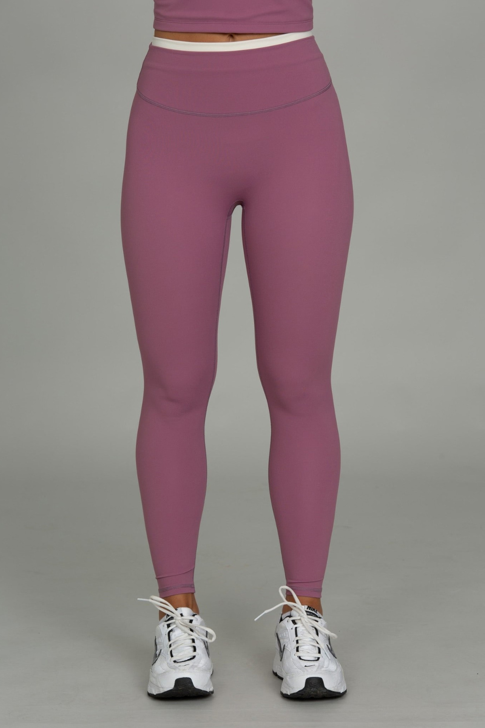 A person from the waist down wearing high-waisted mauve-purple leggings and white athletic sneakers, standing against a plain light gray studio background.