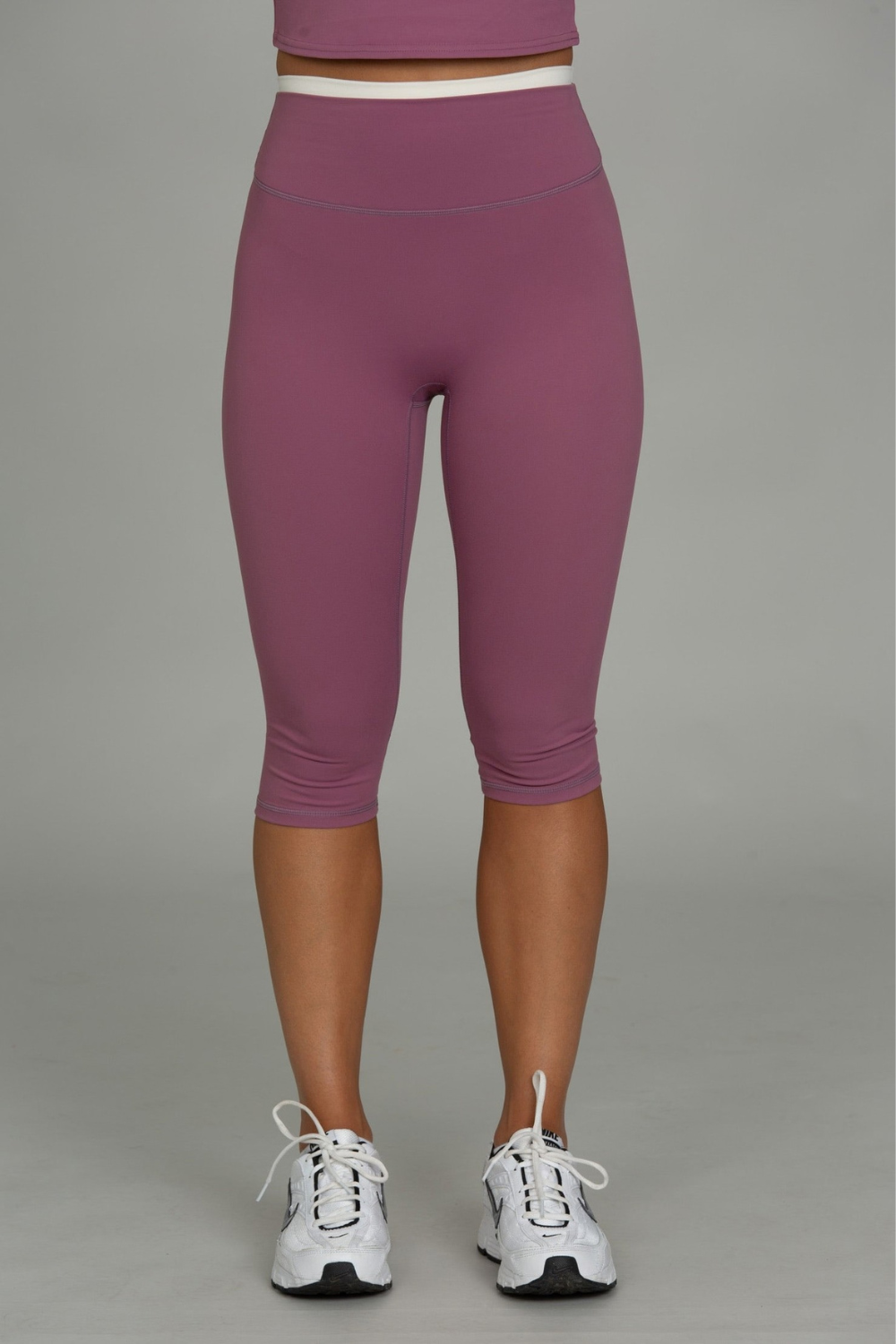 A person from the waist down wearing high-waisted, capri-length athletic leggings in a dusty mauve-purple color, paired with white sneakers, against a solid gray studio background.