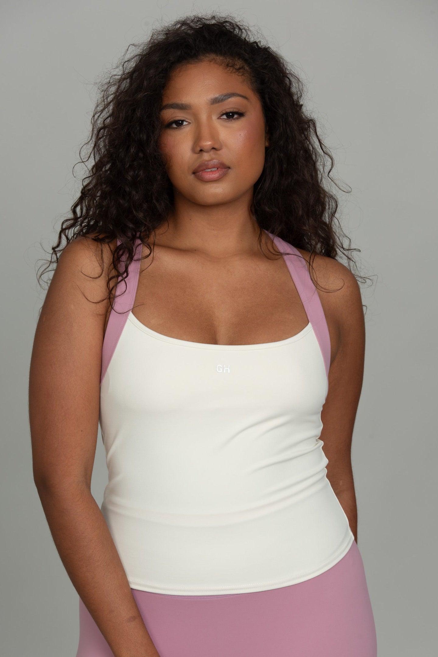 A woman modeling a white scoop-neck tank top with light pink shoulder straps, paired with matching pink leggings, against a solid gray studio background.