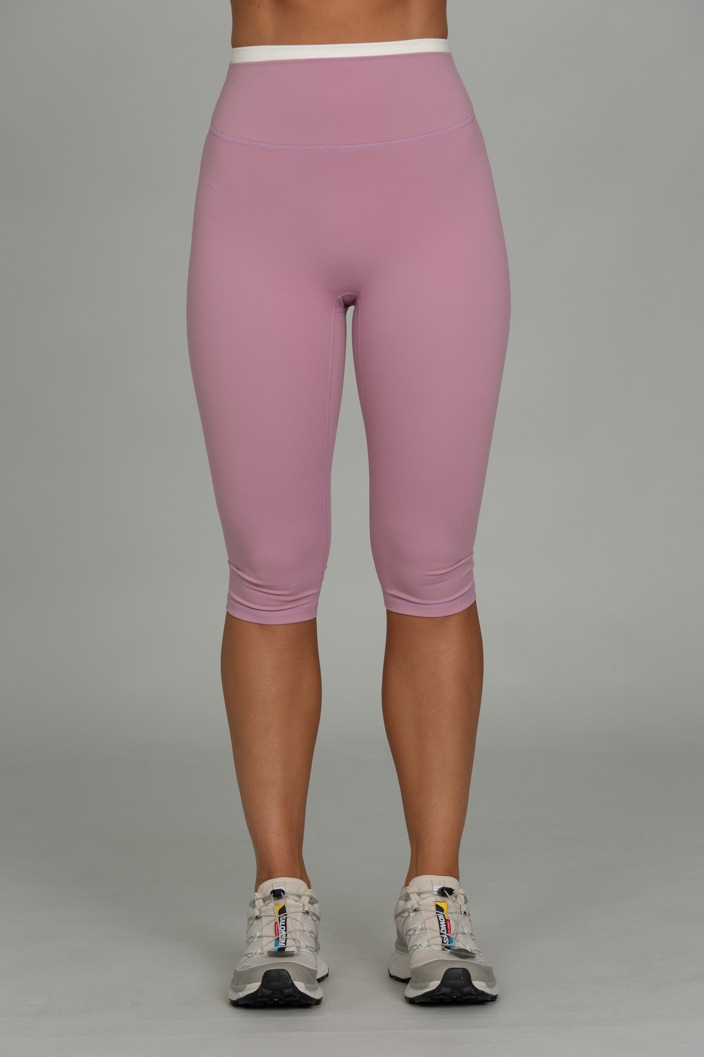 A person shown from the waist down wearing fitted capri leggings in a dusty pink color, paired with gray and white athletic sneakers, against a solid gray studio background.
