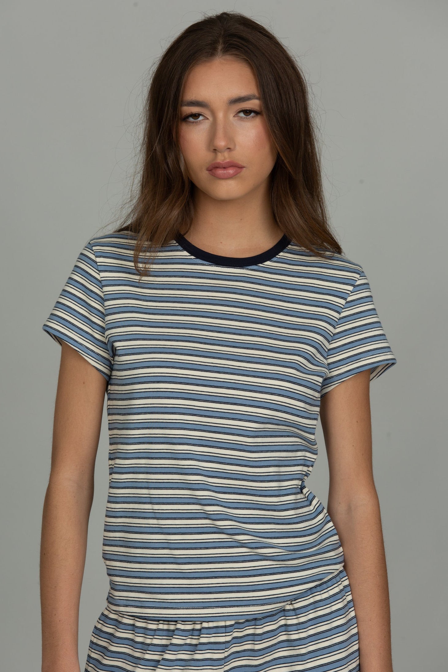 A woman modeling a short-sleeve crewneck t-shirt with horizontal navy, blue, and beige stripes, paired with matching striped bottoms, against a solid gray studio background.