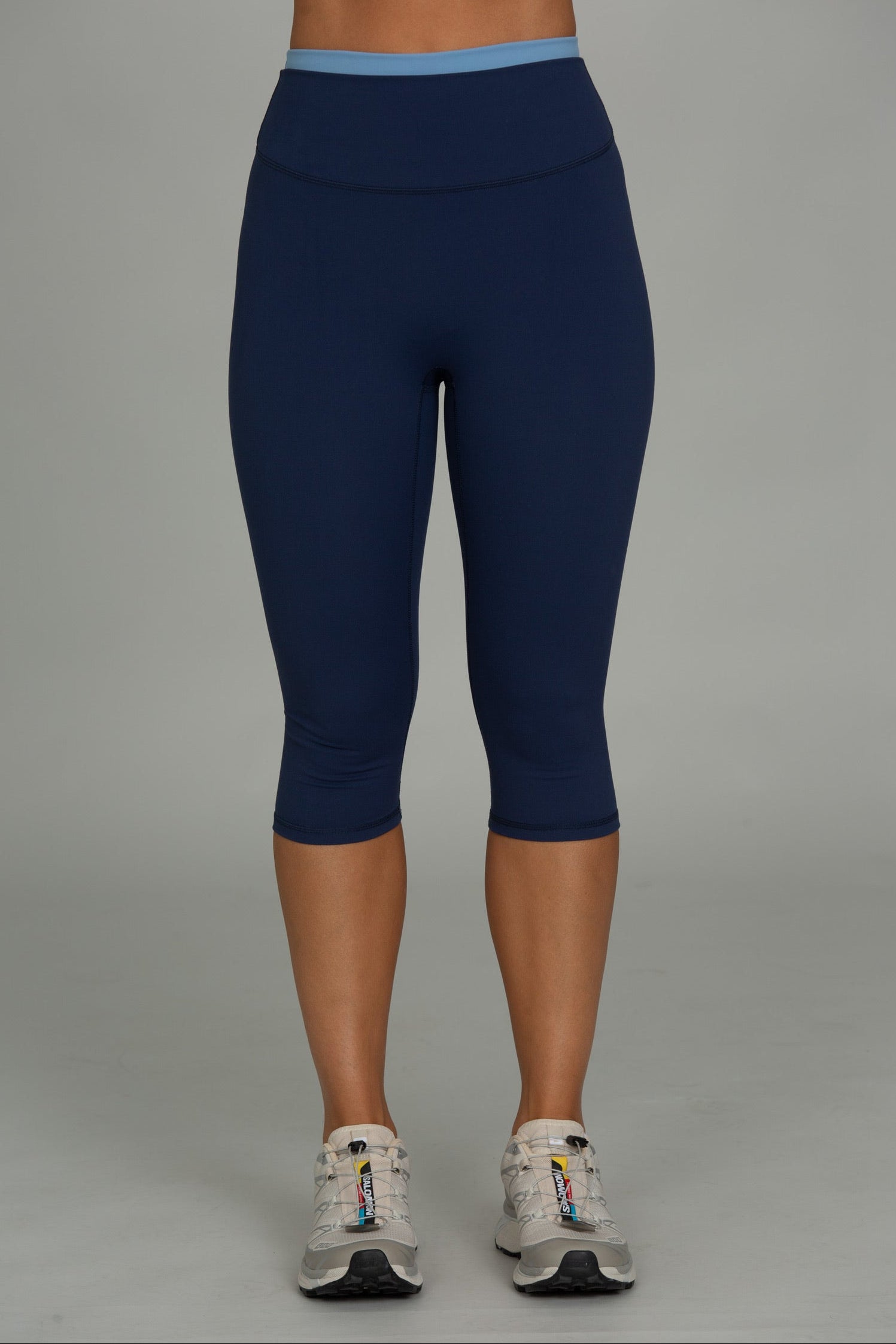 A person from the waist down wearing navy blue high-waisted capri leggings with a light blue waistband, paired with white athletic sneakers, against a solid gray studio background.