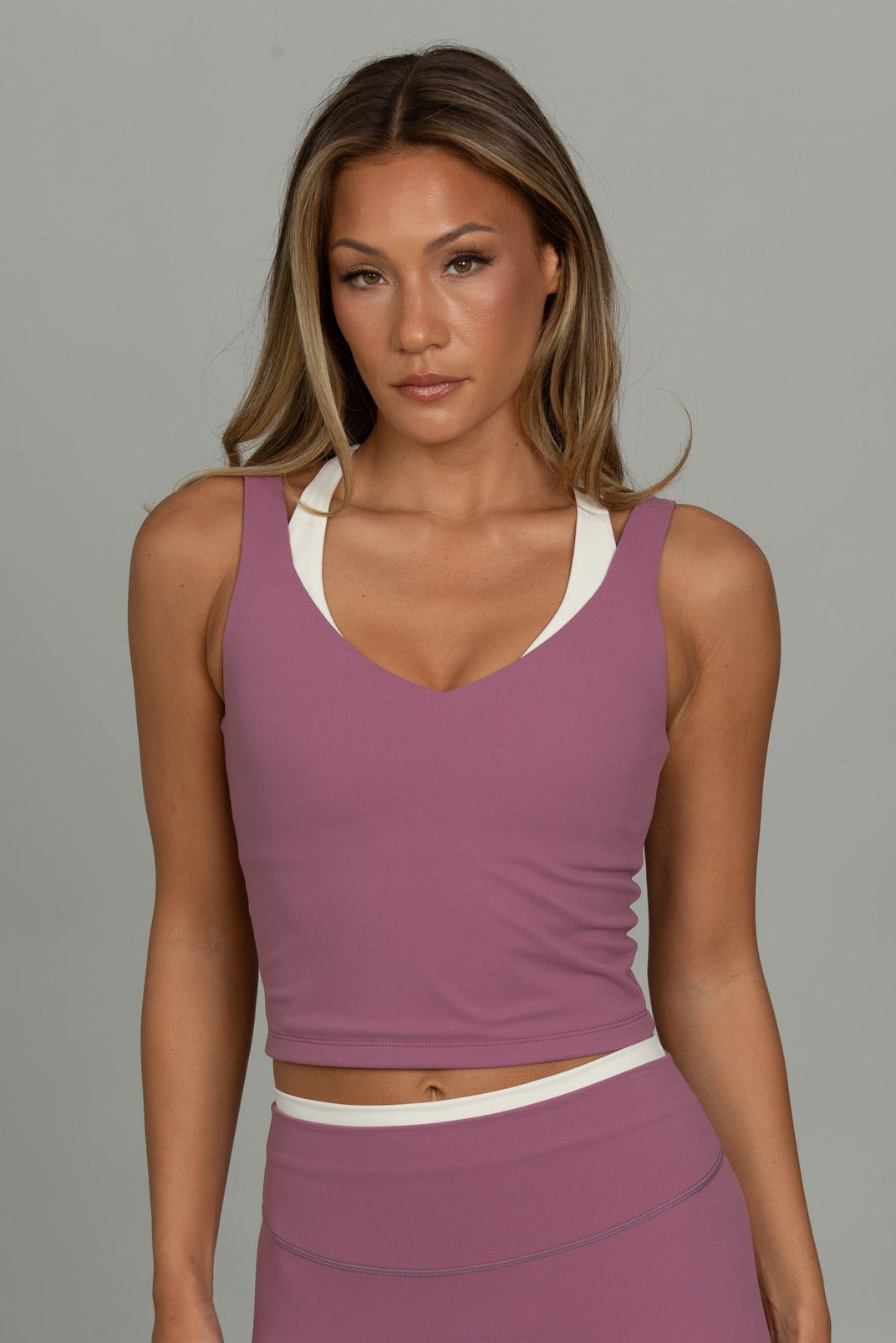 A woman modeling a matching mauve activewear set consisting of a cropped tank top and high-waisted leggings, featuring a contrasting white inner neckline and waistband, set against a solid gray studio background.