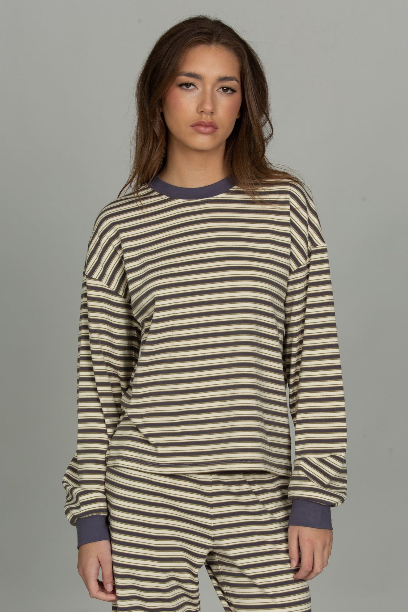 A woman modeling a matching two-piece set with horizontal stripes in beige and dark brown, featuring solid dark purple ribbed trim on the crew neck, cuffs, and ankles, shot against a solid gray studio background.