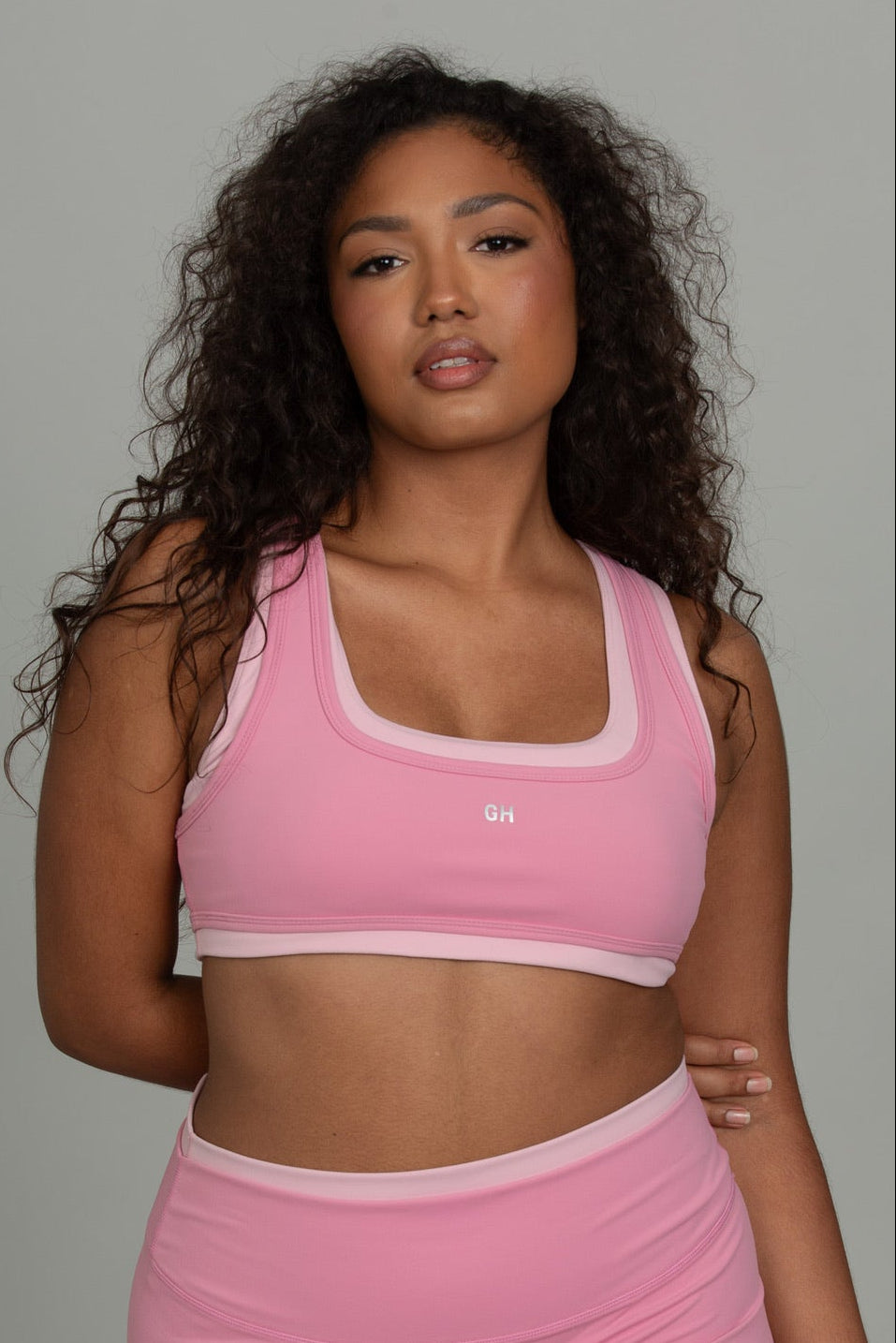 A woman modeling a pink athletic set consisting of a scoop-neck sports bra and matching high-waisted shorts, photographed against a solid gray studio background.