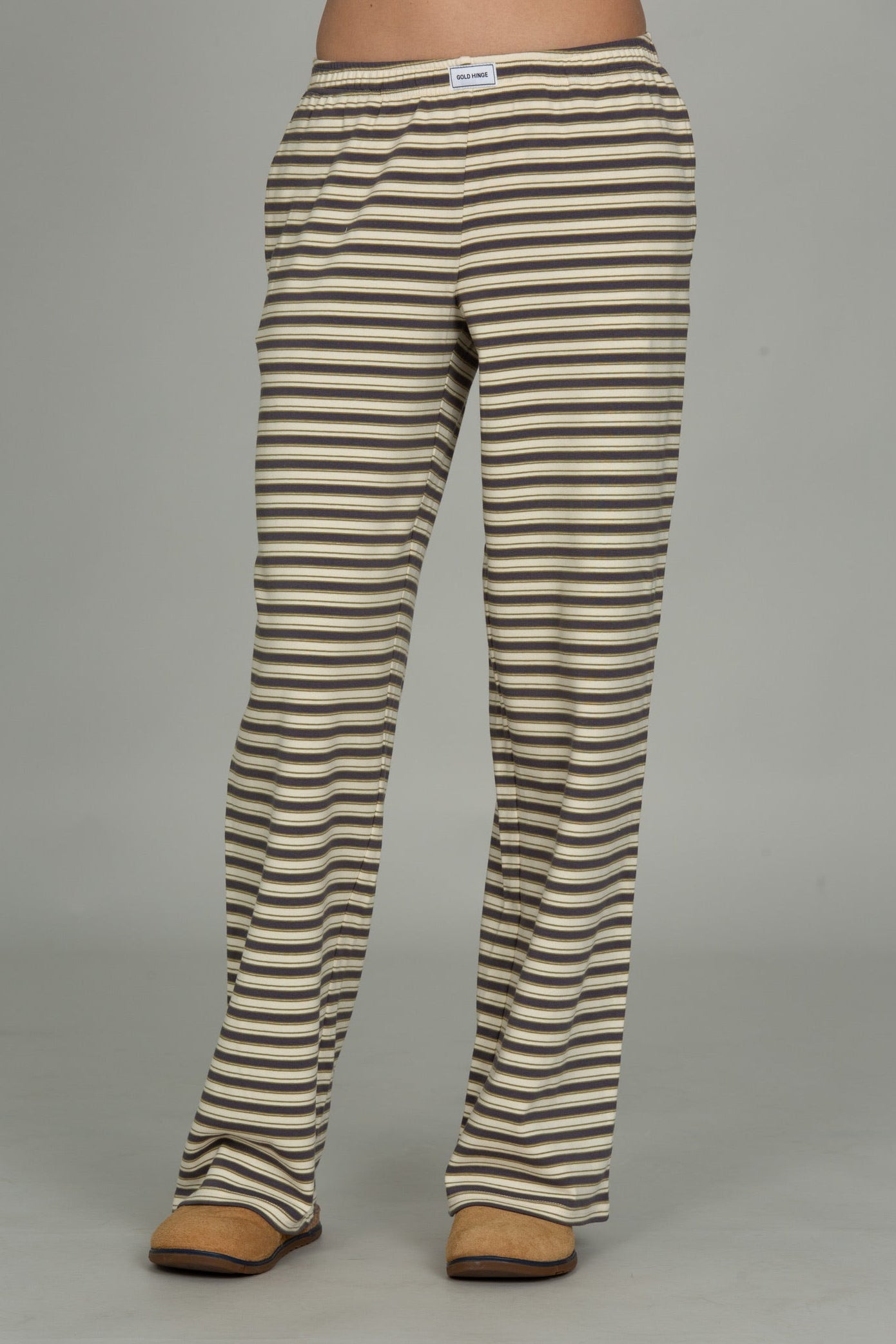A person wearing straight-leg pants with horizontal beige and dark brown stripes, paired with tan shoes, shown from the waist down against a plain gray studio background.