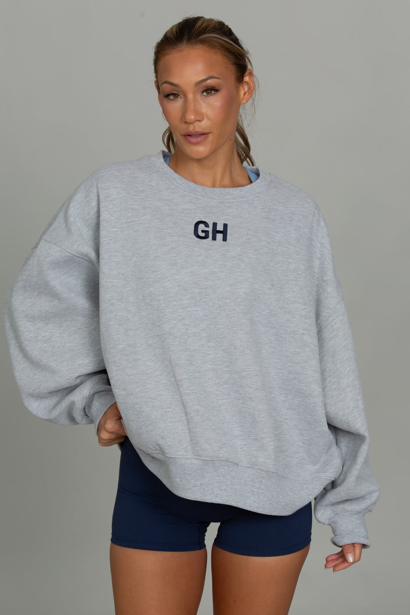 A woman modeling an oversized light gray heather sweatshirt with 'GH' printed in navy on the chest, paired with navy athletic shorts, against a solid gray studio background.