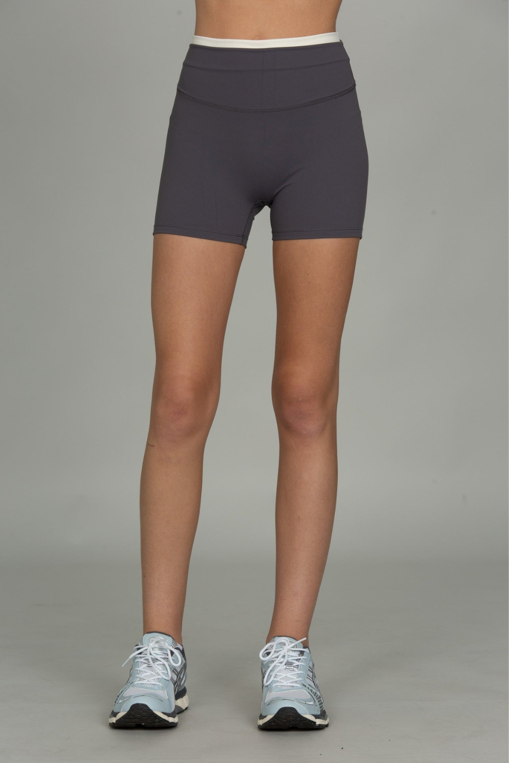 A person from the waist down wearing dark charcoal gray athletic biker shorts with a white waistband, paired with light blue running shoes, against a solid gray studio background.