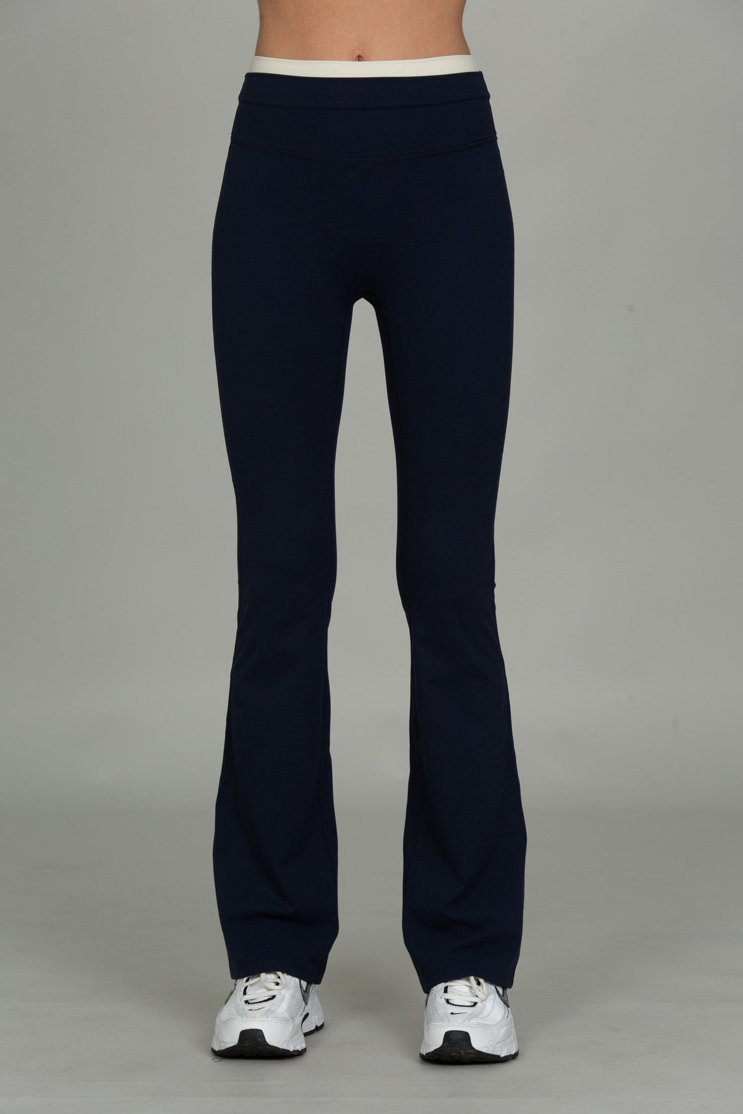 A person from the waist down wearing dark navy flared leggings or bootcut pants with a contrasting white waistband, paired with white sneakers, against a neutral gray studio background.