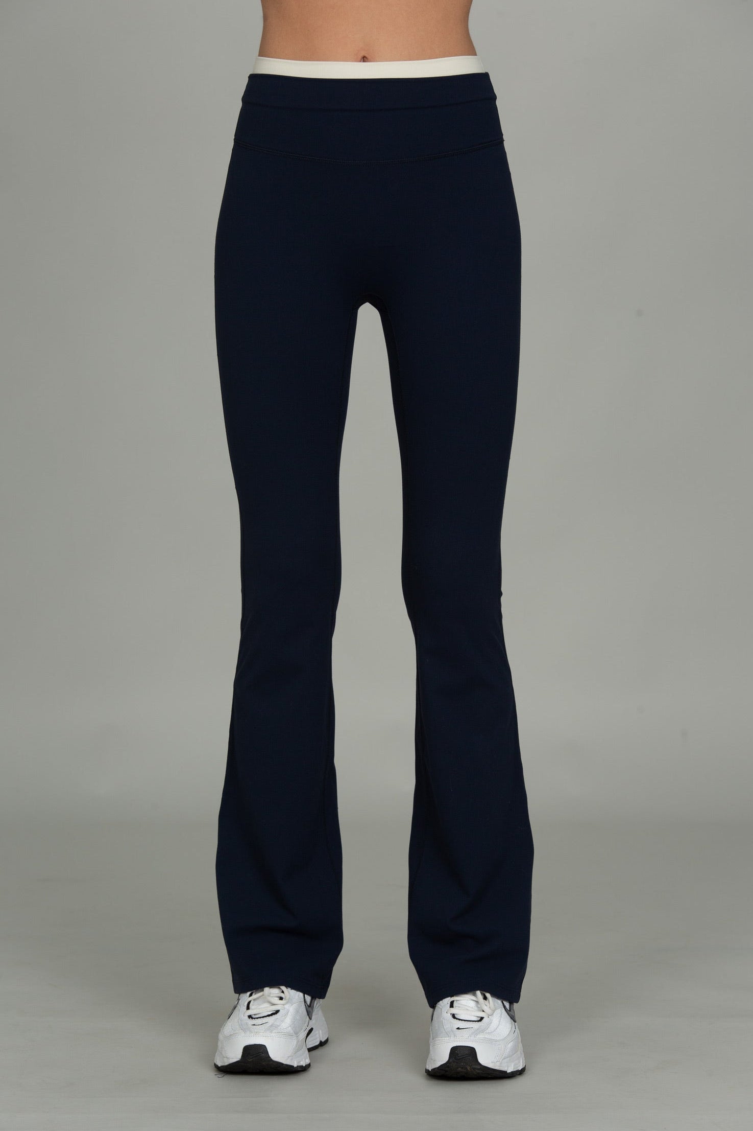 A person from the waist down wearing dark navy flared leggings or bootcut pants with a contrasting white waistband, paired with white sneakers, against a neutral gray studio background.