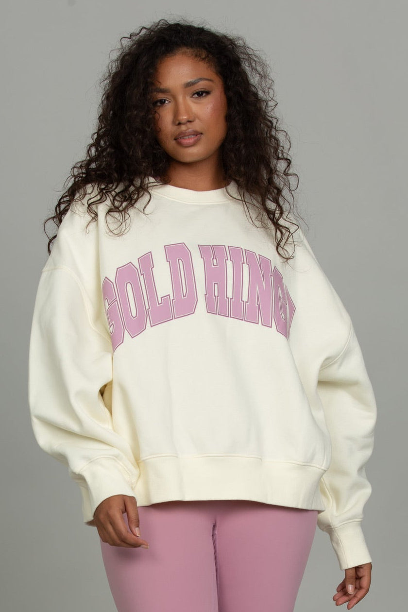 A model wearing a cream-colored oversized sweatshirt with pink 'GOLD HINGE' text on the front, paired with pink capri leggings and white sneakers, standing against a solid gray studio background.
