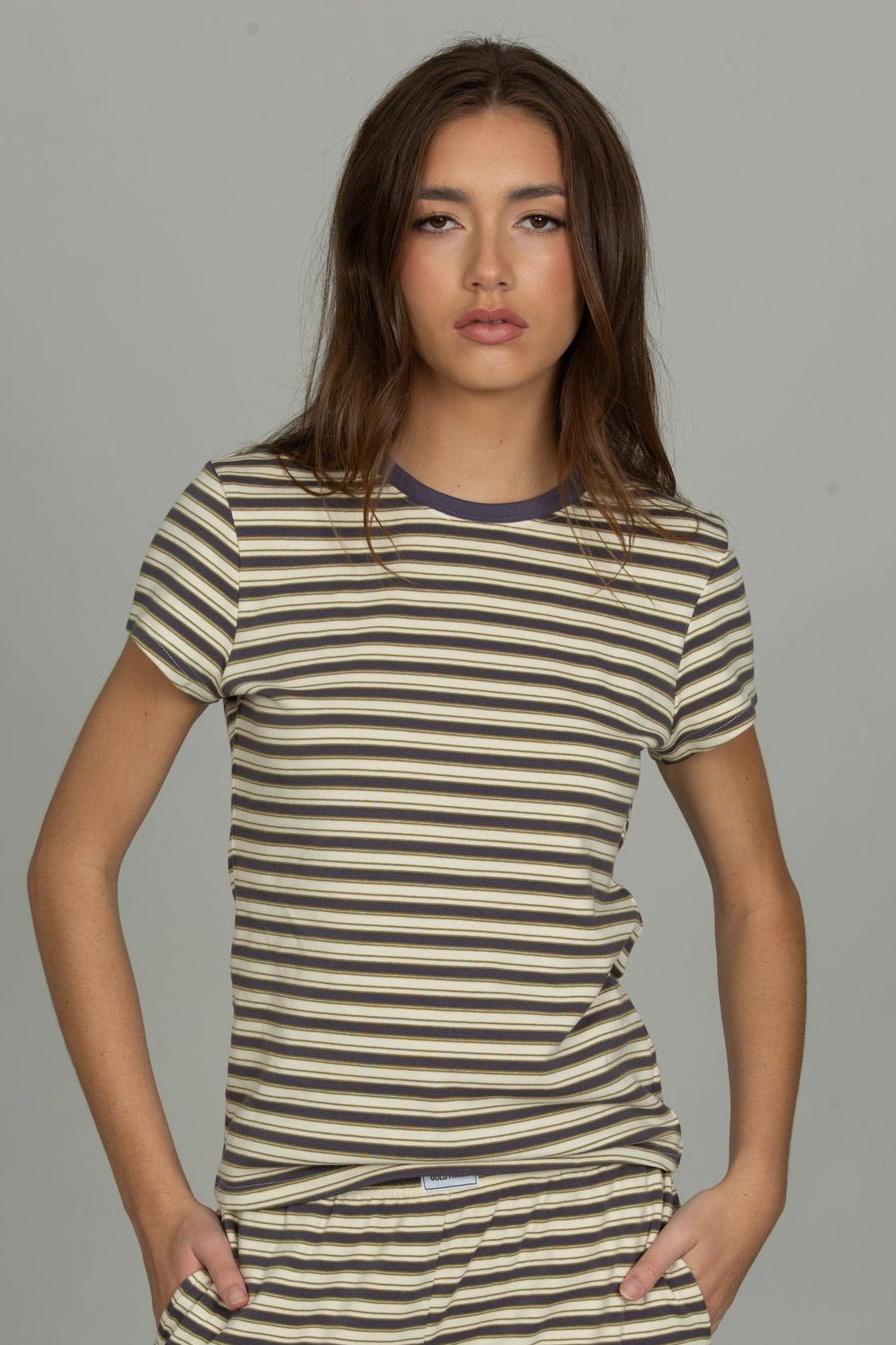 A woman modeling a short-sleeve crewneck t-shirt with horizontal beige and dark brown stripes, featuring a contrasting dark navy collar, set against a solid gray studio background.
