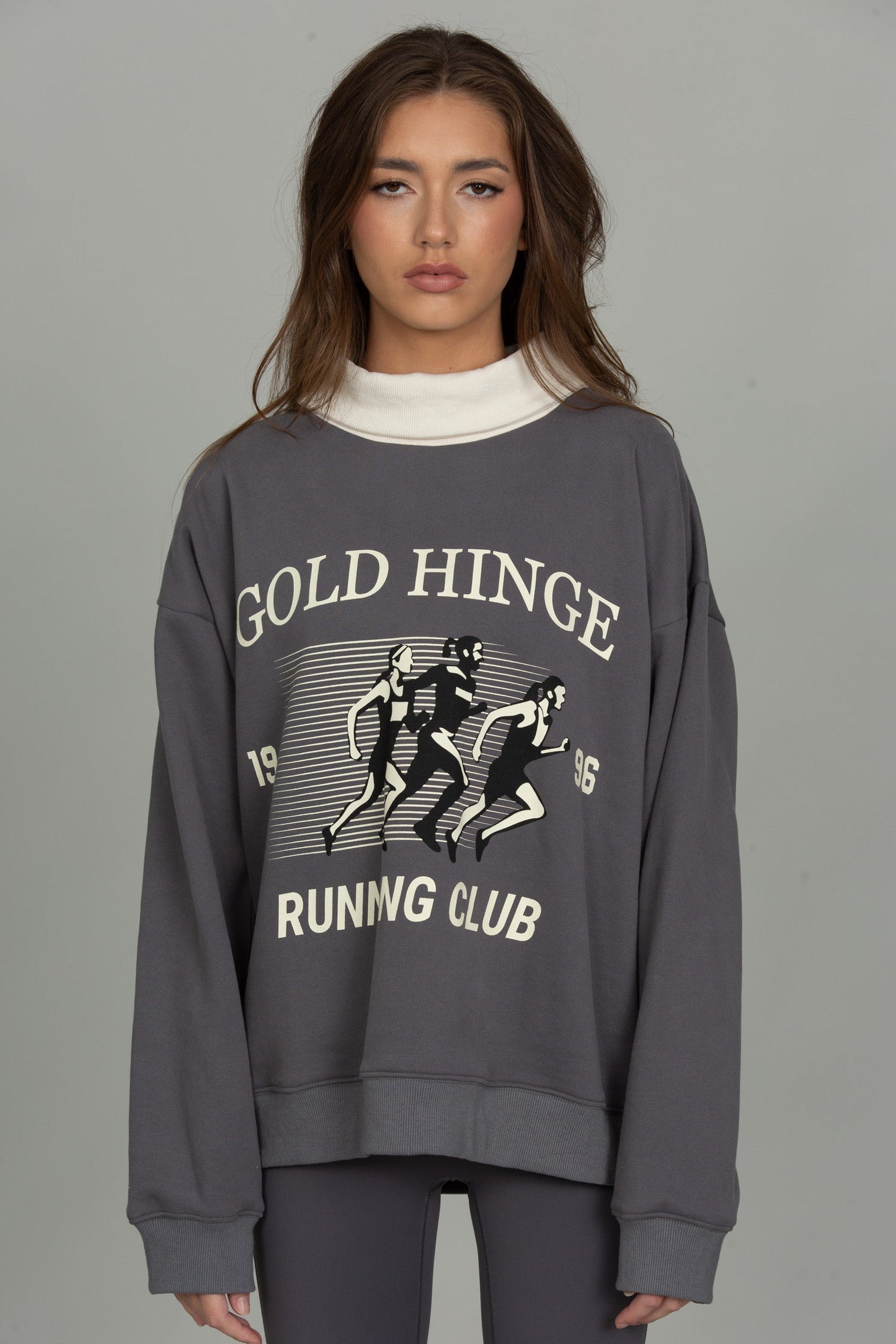 A woman modeling a dark gray crewneck sweatshirt with a vintage-style graphic that reads 'GOLD HINGE RUNNING CLUB' in white and black, layered over a white mock neck.