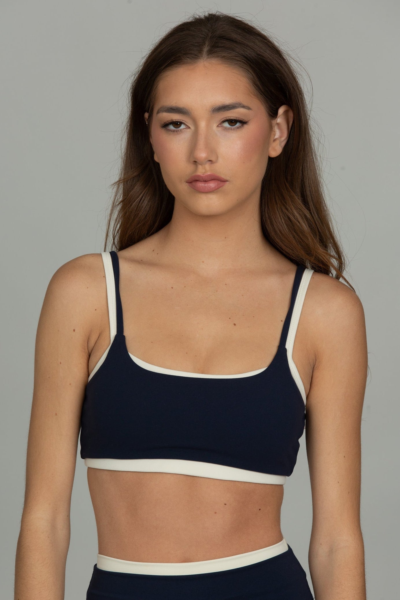 A woman modeling a navy blue sports bra with contrasting white trim, paired with matching navy high-waisted shorts, against a solid gray studio background.
