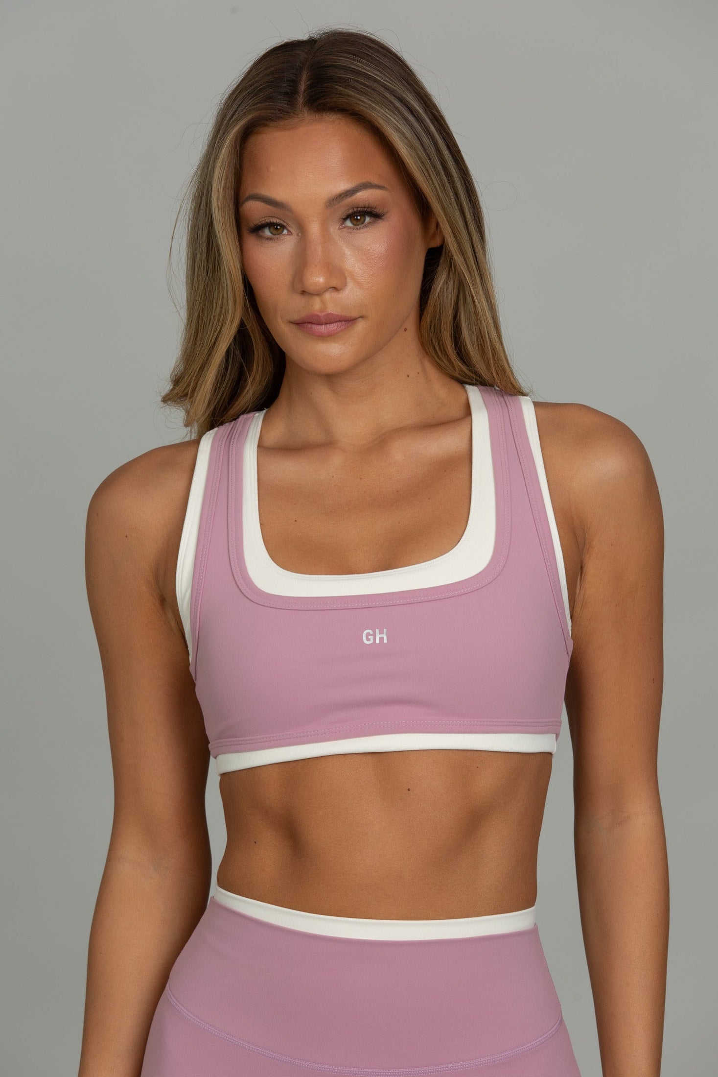A woman modeling a dusty pink athletic sports bra with white trim and a matching high-waisted athletic bottom against a solid gray studio background.
