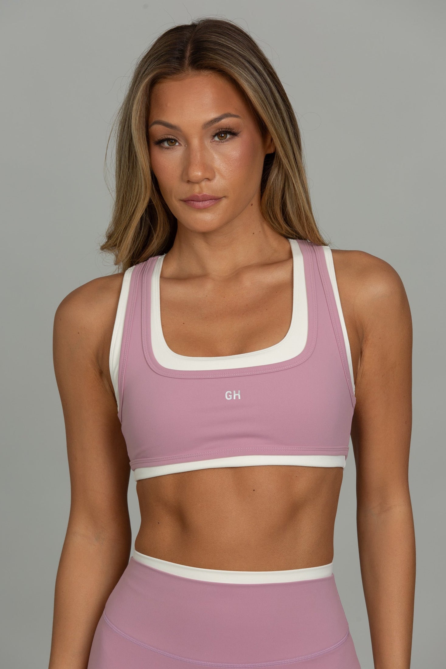 A woman modeling a dusty pink athletic sports bra with white trim and a matching high-waisted athletic bottom against a solid gray studio background.