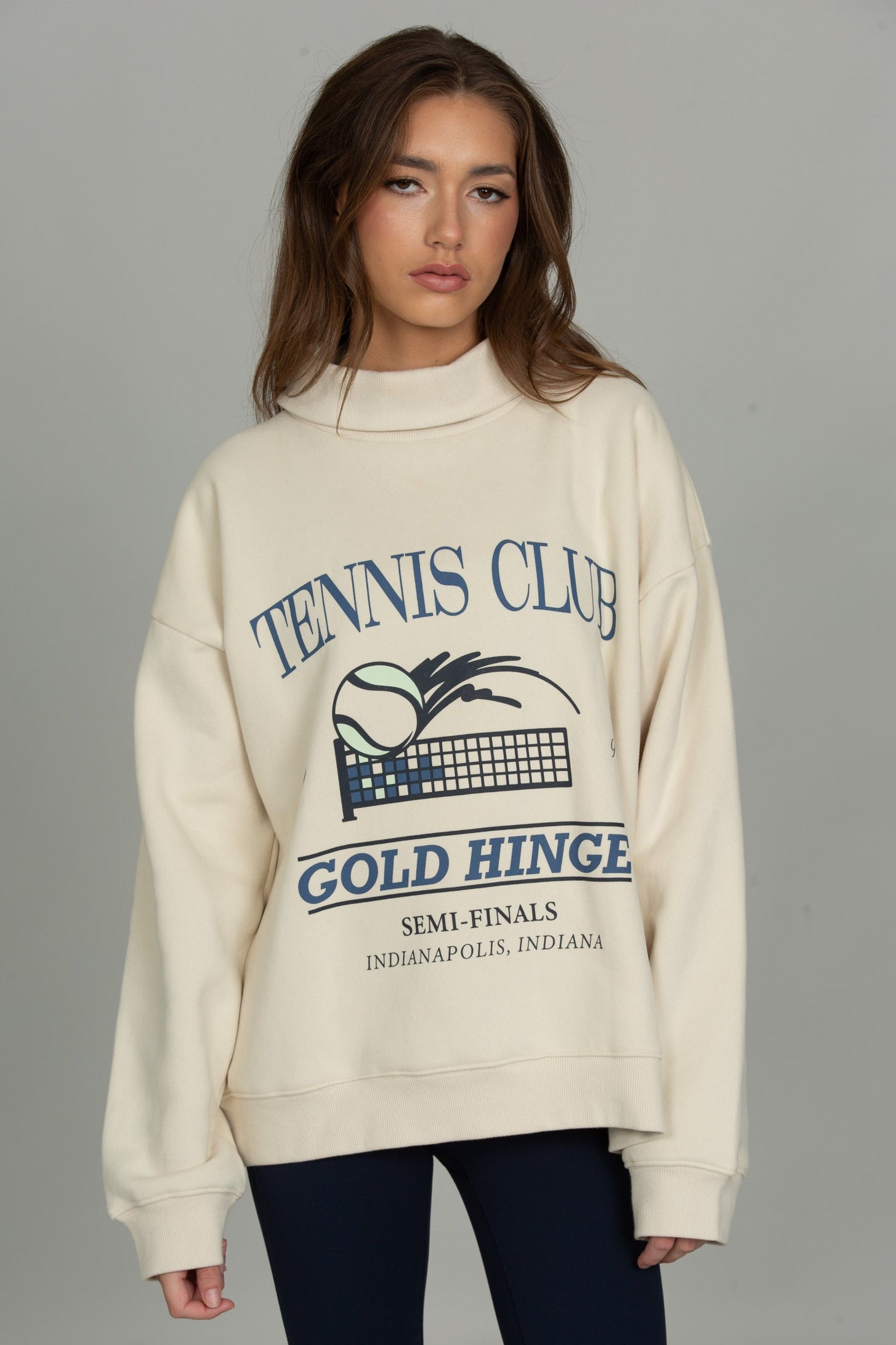 A woman modeling an oversized cream sweatshirt with a navy blue 'TENNIS CLUB GOLD HINGE' graphic on the front, paired with dark leggings, against a solid gray studio background.