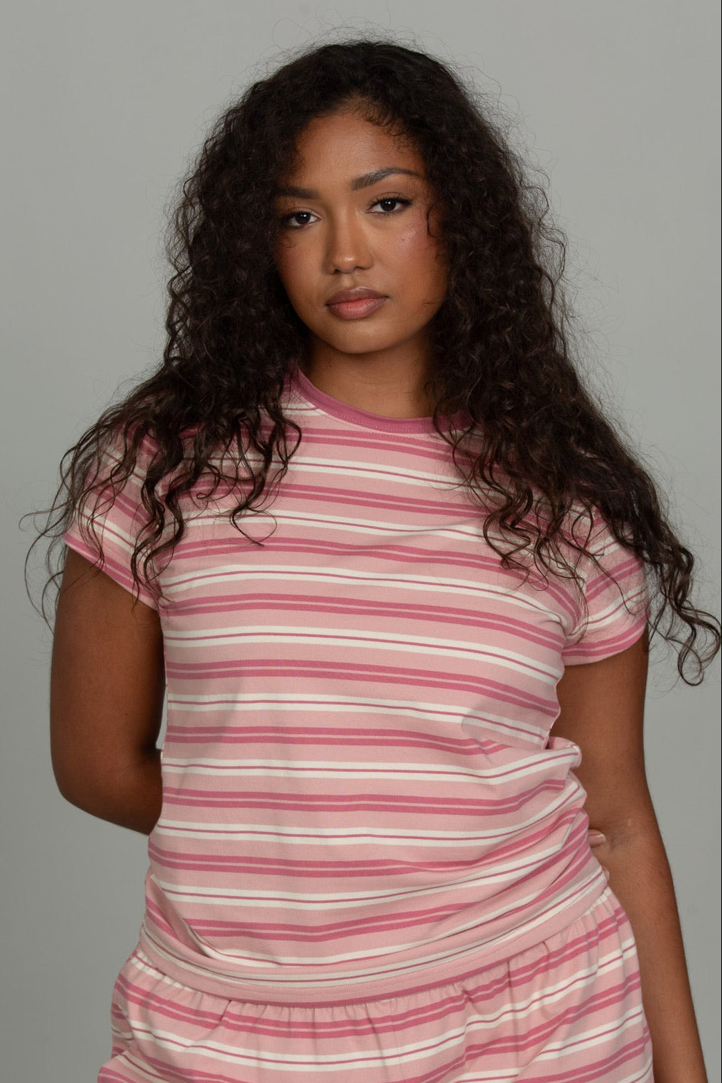 A woman modeling a matching two-piece loungewear set consisting of a short-sleeve crewneck t-shirt and shorts, both featuring horizontal pink and white stripes, against a solid gray studio background.