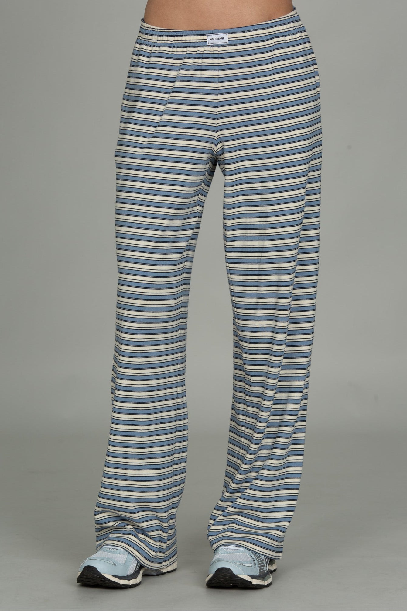 A person from the waist down wearing striped lounge pants in blue, gray, and white, paired with light blue sneakers, against a solid gray studio background.