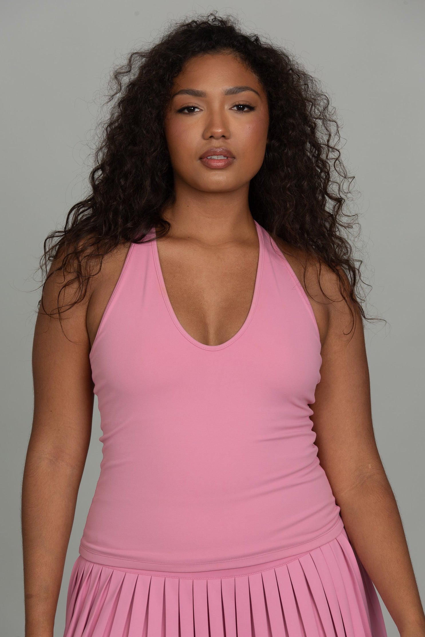 A person wearing a pink halter-neck top and matching pleated skirt, photographed against a solid gray studio background.