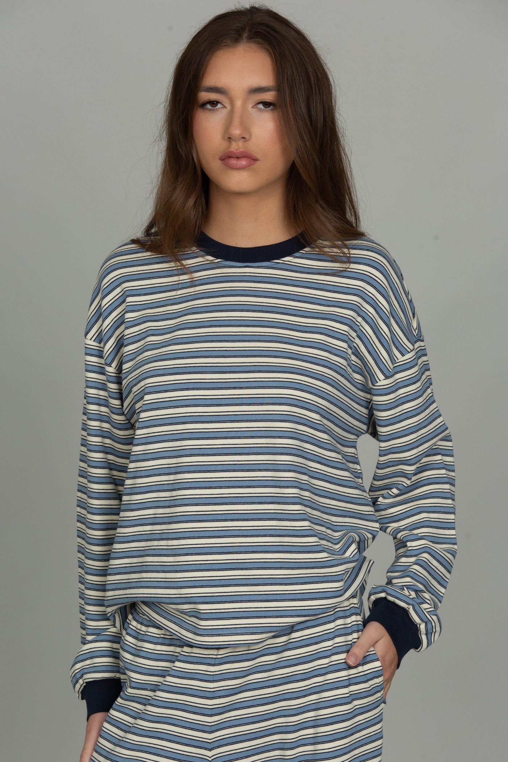 A woman modeling a matching striped lounge set consisting of a long-sleeve top and pants in blue, beige, and white horizontal stripes with dark navy cuffs and collar.
