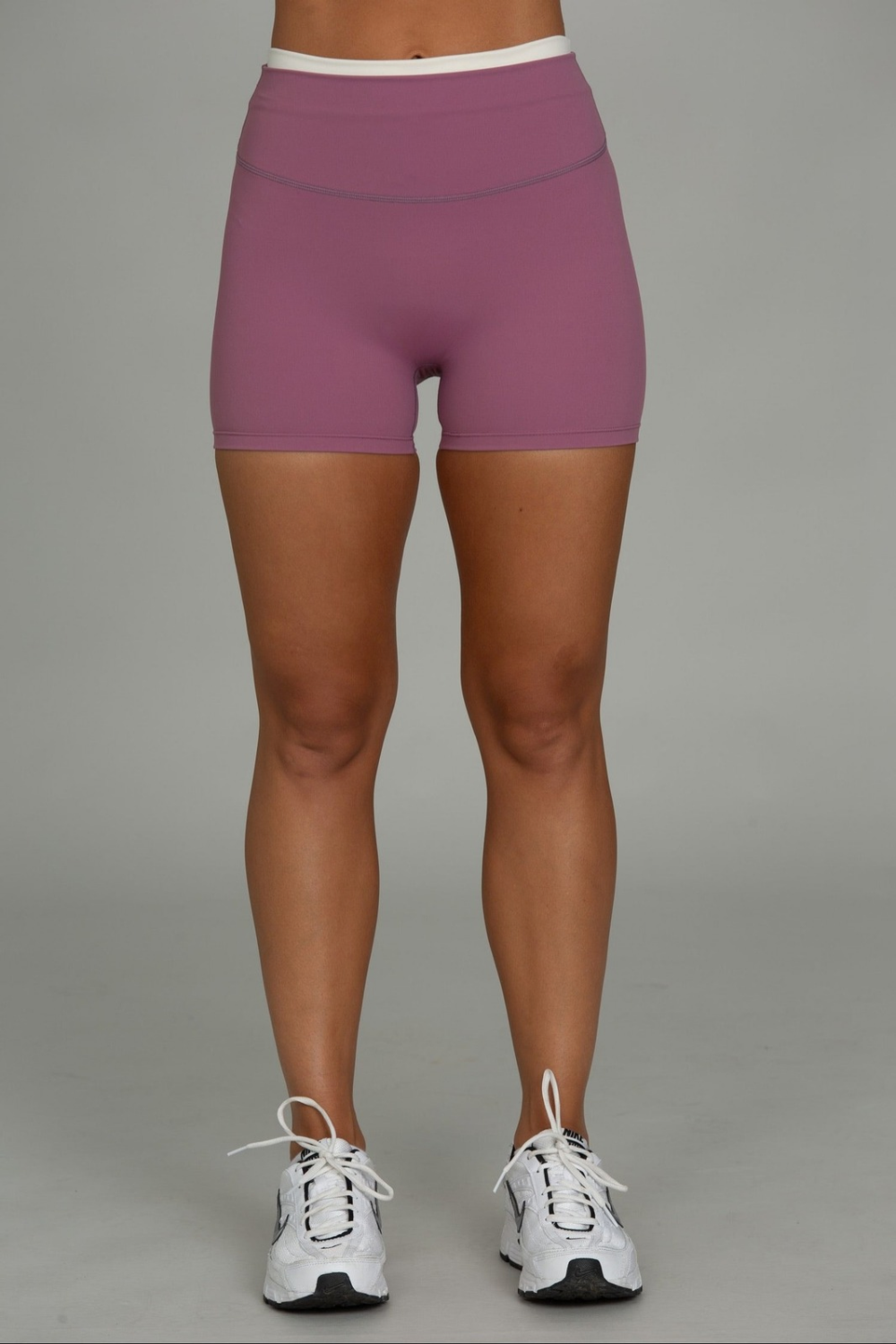 A person from the waist down wearing high-waisted, form-fitting athletic shorts in a dusty mauve-purple color, paired with white sneakers, against a solid gray studio background.