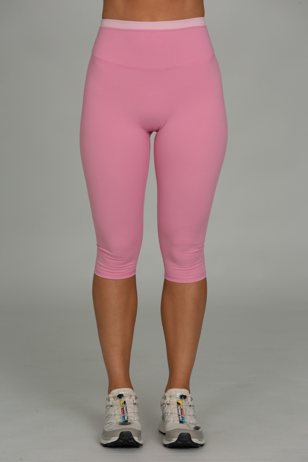 A person from the waist down wearing pink capri leggings and white athletic sneakers, standing against a plain light gray studio background.
