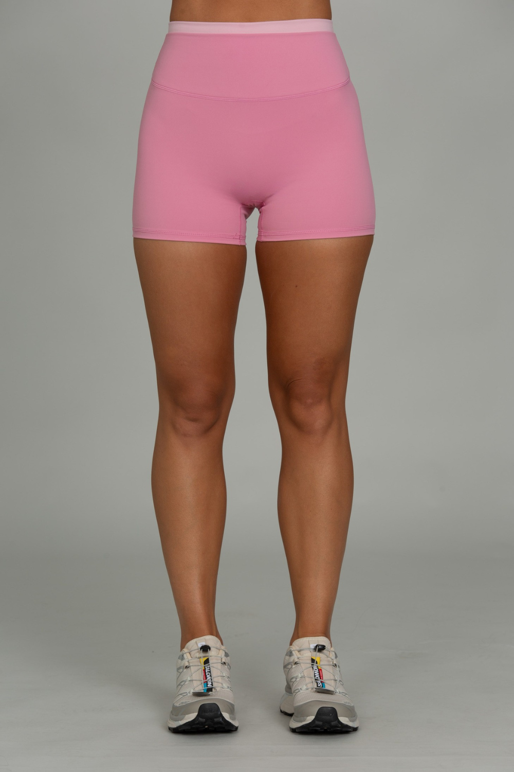 A person from the waist down wearing bright pink athletic shorts and white running shoes, standing against a plain light gray studio background.