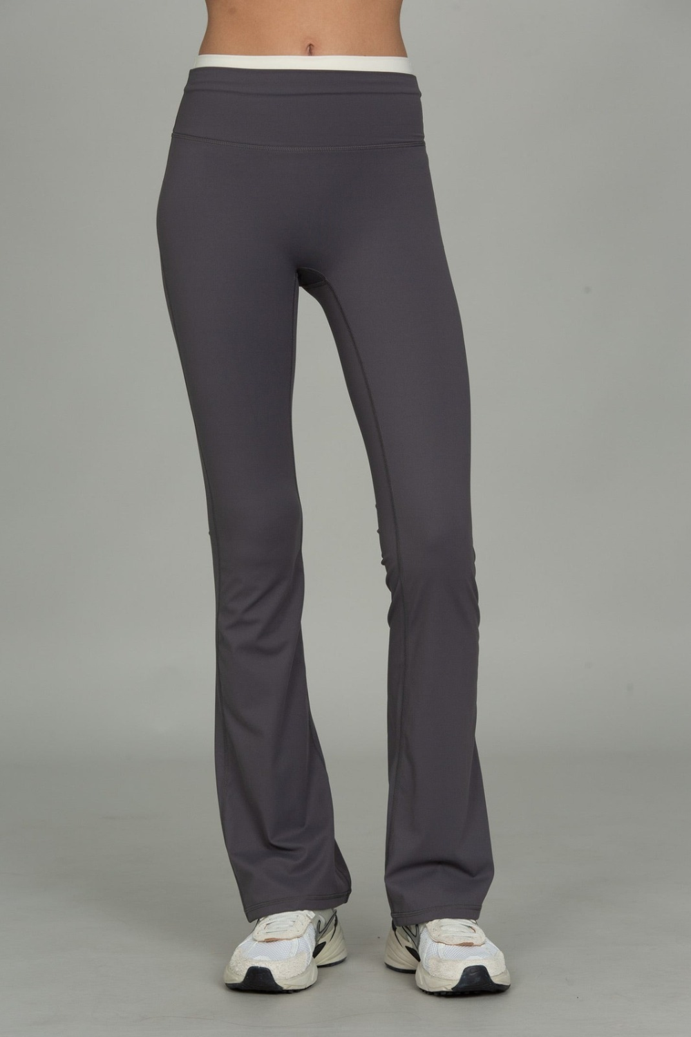 A person from the waist down wearing dark gray flared leggings and white sneakers, standing against a plain light gray studio background.