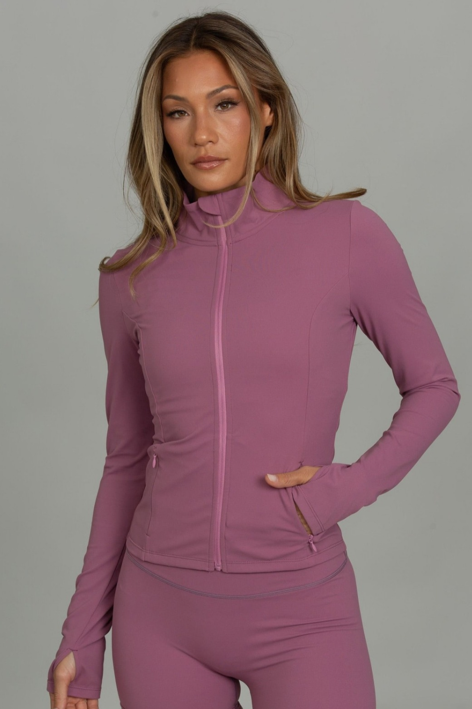 A woman modeling a matching athletic set consisting of a long-sleeve zip-up jacket and fitted shorts in a dusty mauve color, set against a solid gray studio background.